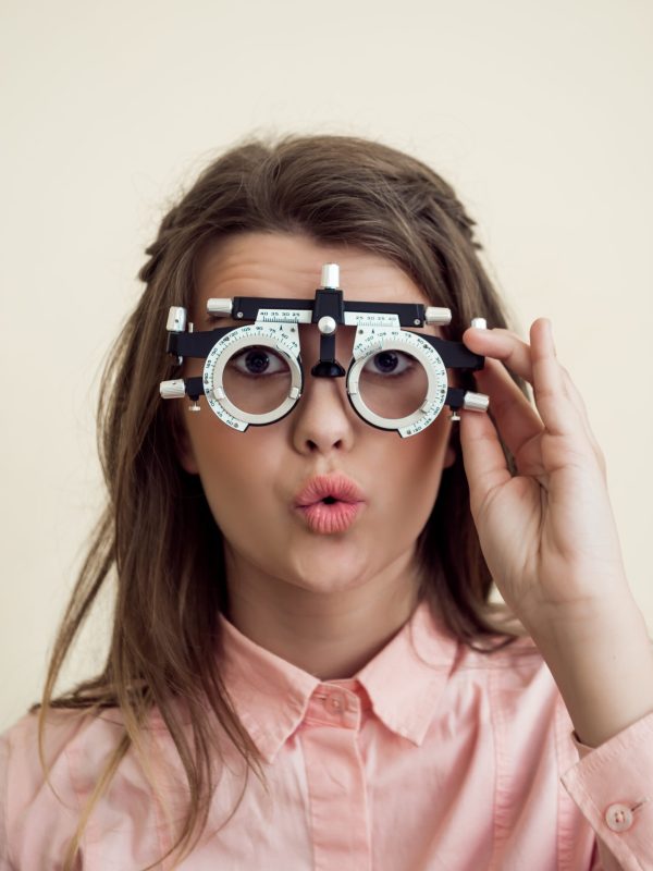 Horizontal shot of excited cute european brunette checking vision with phoropter, being interested in how it works, waiting for optometrist to prescribe appropriate glasses. Eyecare concept